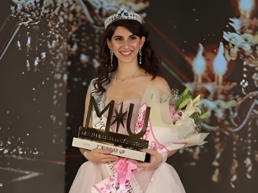 Simran standing on a pageant stage in a pink evening gown, holding a trophy and a bouquet of flowers.